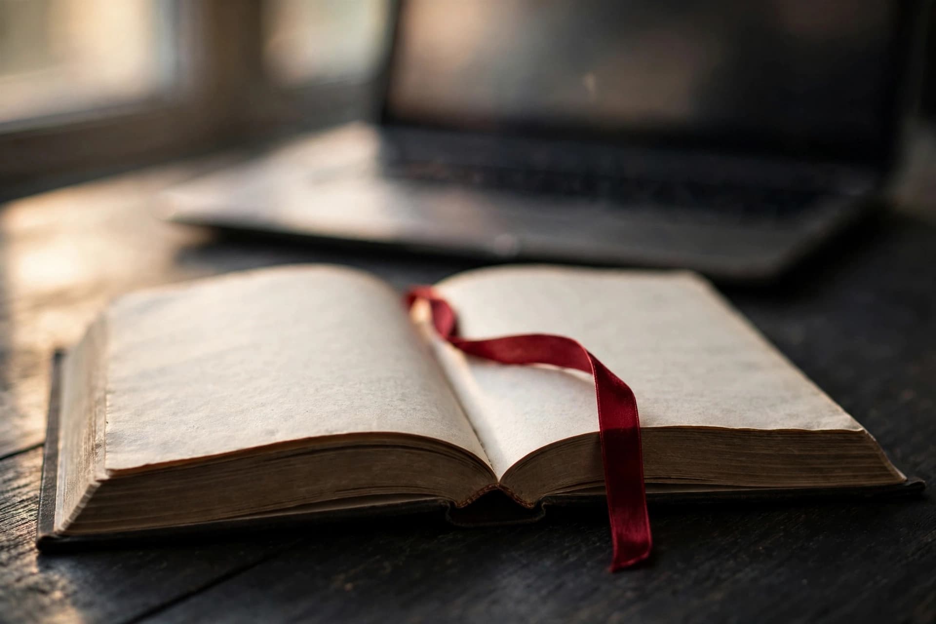 Open book on a desk with a crimson-red ribbon bookmark beside a laptop showing a PDF contents page